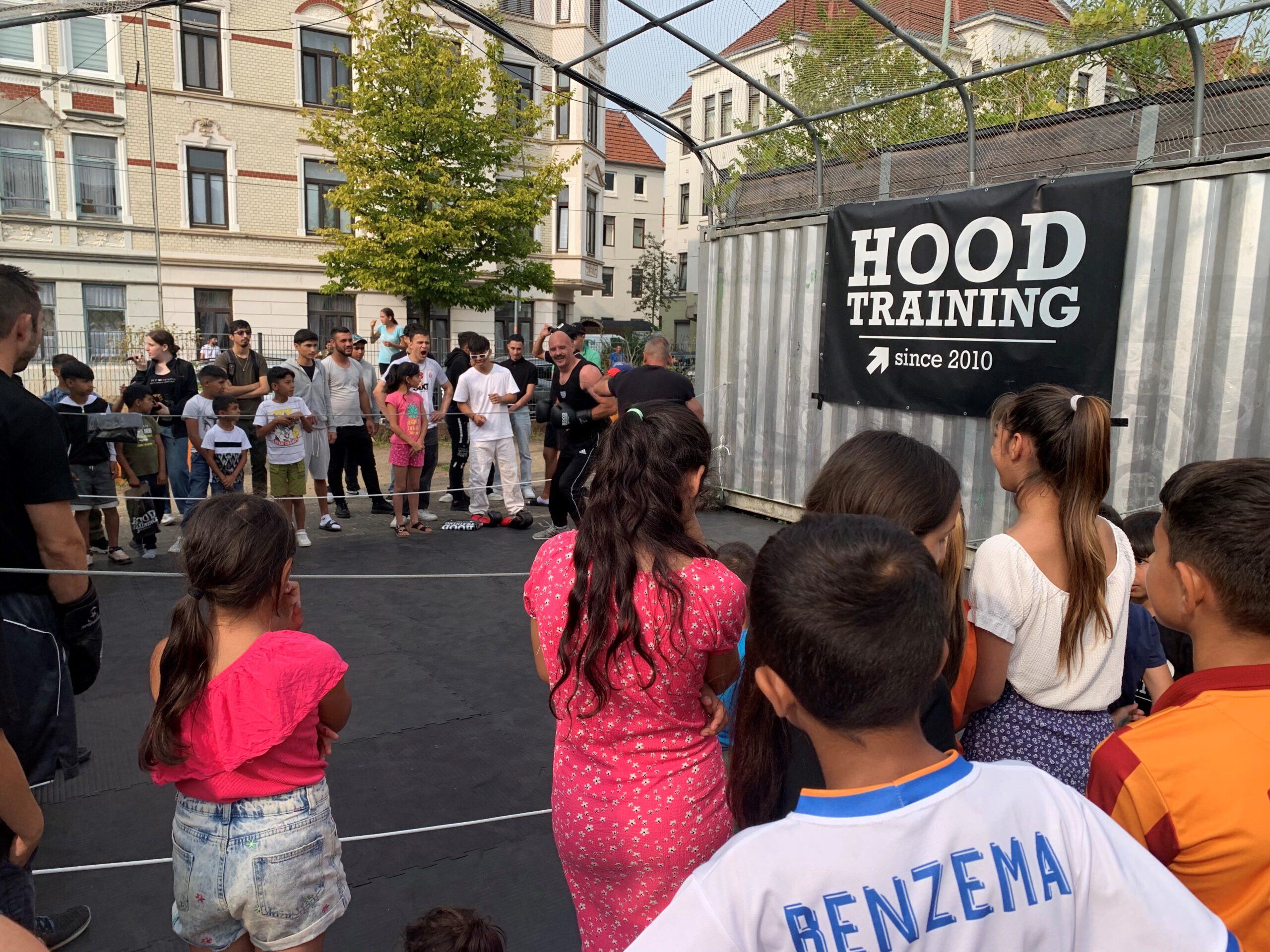 Viele Menschen jeden Alters stehen um eine mit Matten ausgelegte Fläche, die von der einen Seite von einem Container begrenzt wird, an welchem ein schwarzes Banner mit weißer Schrift und 