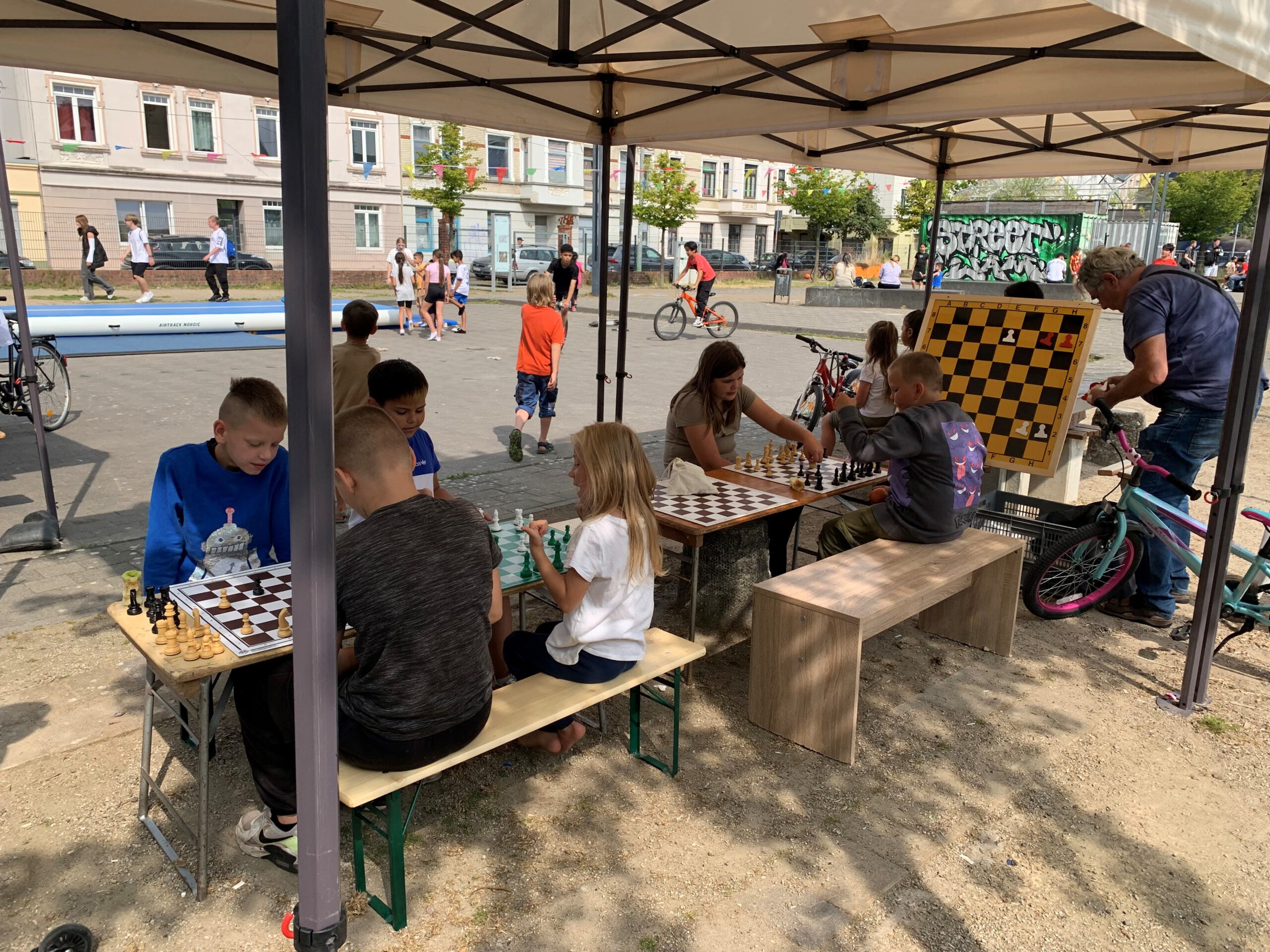 Unter zwei Pavillons sind kleine Bierzelttische und -bänke für Kinder aufgebaut, an denen Kinder vor Schachbrettern sitzen. Im Hintergrund laufen einzelne Kinder herum. An der Seite steht ein großes Schach-Demoboard.