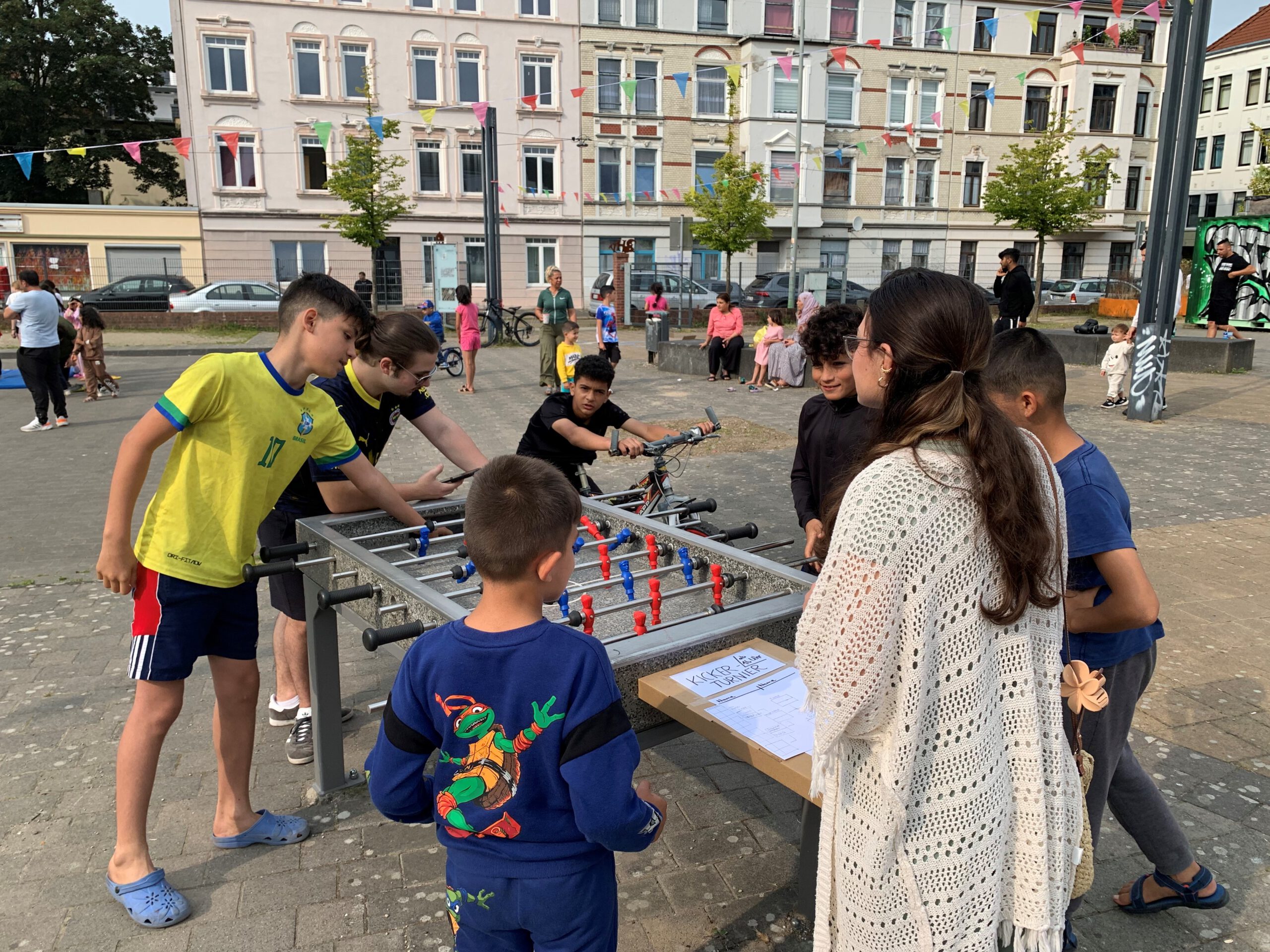 Um einen Tischkicker stehen Kinder und Jugendliche herum. Im Hintergrund sind vor einer Altbaufassade weitere Kinder und eine bunte Wimpelkette.