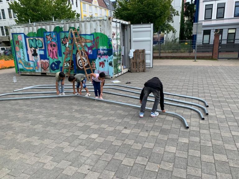 Vier Kinder sind dabei eine Stange des Gerüsts für das „Leher Pergolinchen“ anzuheben. Im Hintergrund sieht man eine Leiter und einen der zwei Container, auf dem das Gerüst befestigt werden soll.