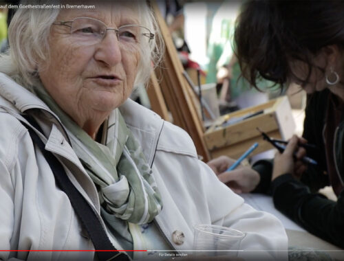 Videovorschaubild für das Goethestraßenfest. Ein ältere Frau mit einer Brille spricht. Im Hintergrund sieht man eine dunkelhaarige Frau mit Ohrringen, die etwas zeichnet.