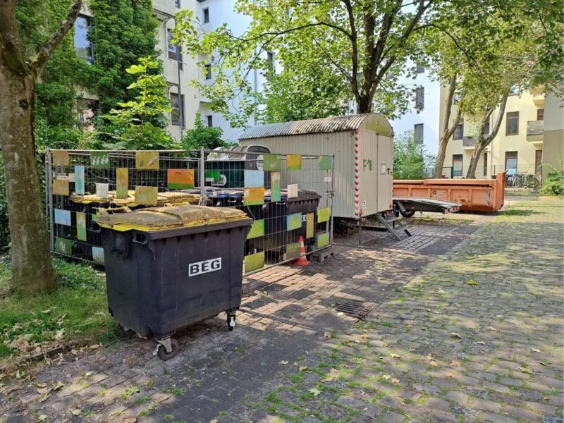 Eingezäunte Müll-Container in der Frenssenstraße. Hinter den Containern steht ein Bauwagen für die Angestellten.