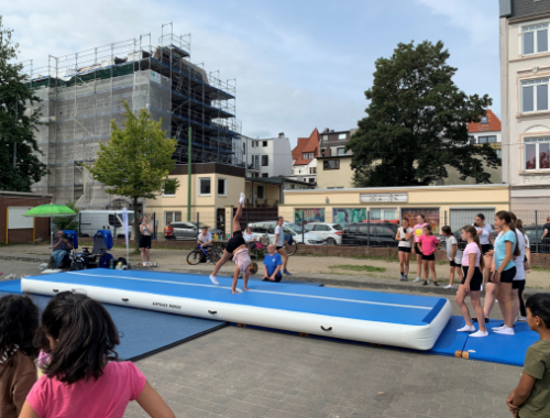 Blauweiße Airtrackmatte liegt draußen auf einer Fläche. Viele Kinder darum versammelt, eine Turnerin befindet sich auf der Matte und macht einen Überschlag.