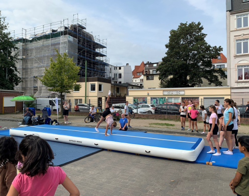 Blauweiße Airtrackmatte liegt draußen auf einer Fläche. Viele Kinder darum versammelt, eine Turnerin befindet sich auf der Matte und macht einen Überschlag.