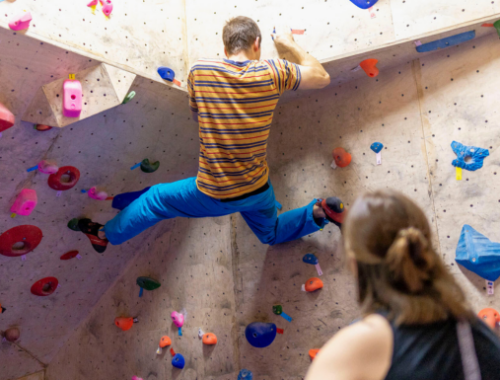 Männliche Person bouldert an einer Boulderwand. Im Vordergrund befindet sich eine weibliche Person, die ihm dabei zuschaut.