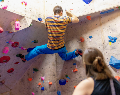 Männliche Person bouldert an einer Boulderwand. Im Vordergrund befindet sich eine weibliche Person, die ihm dabei zuschaut.