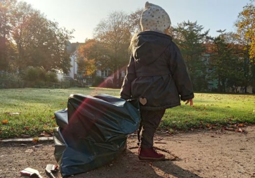 Kleines Mädchen läuft mit einem großen Müllsack durch den Stadtpark Lehe. Die Sonne scheint und der Morgentau liegt noch auf dem Gras im Hintergrund.