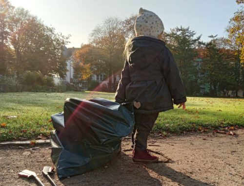 Kleines Mädchen läuft mit einem großen Müllsack durch den Stadtpark Lehe. Die Sonne scheint und der Morgentau liegt noch auf dem Gras im Hintergrund.