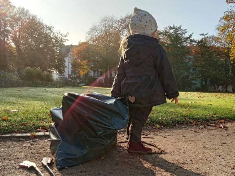 Kleines Mädchen läuft mit einem großen Müllsack durch den Stadtpark Lehe. Die Sonne scheint und der Morgentau liegt noch auf dem Gras im Hintergrund.