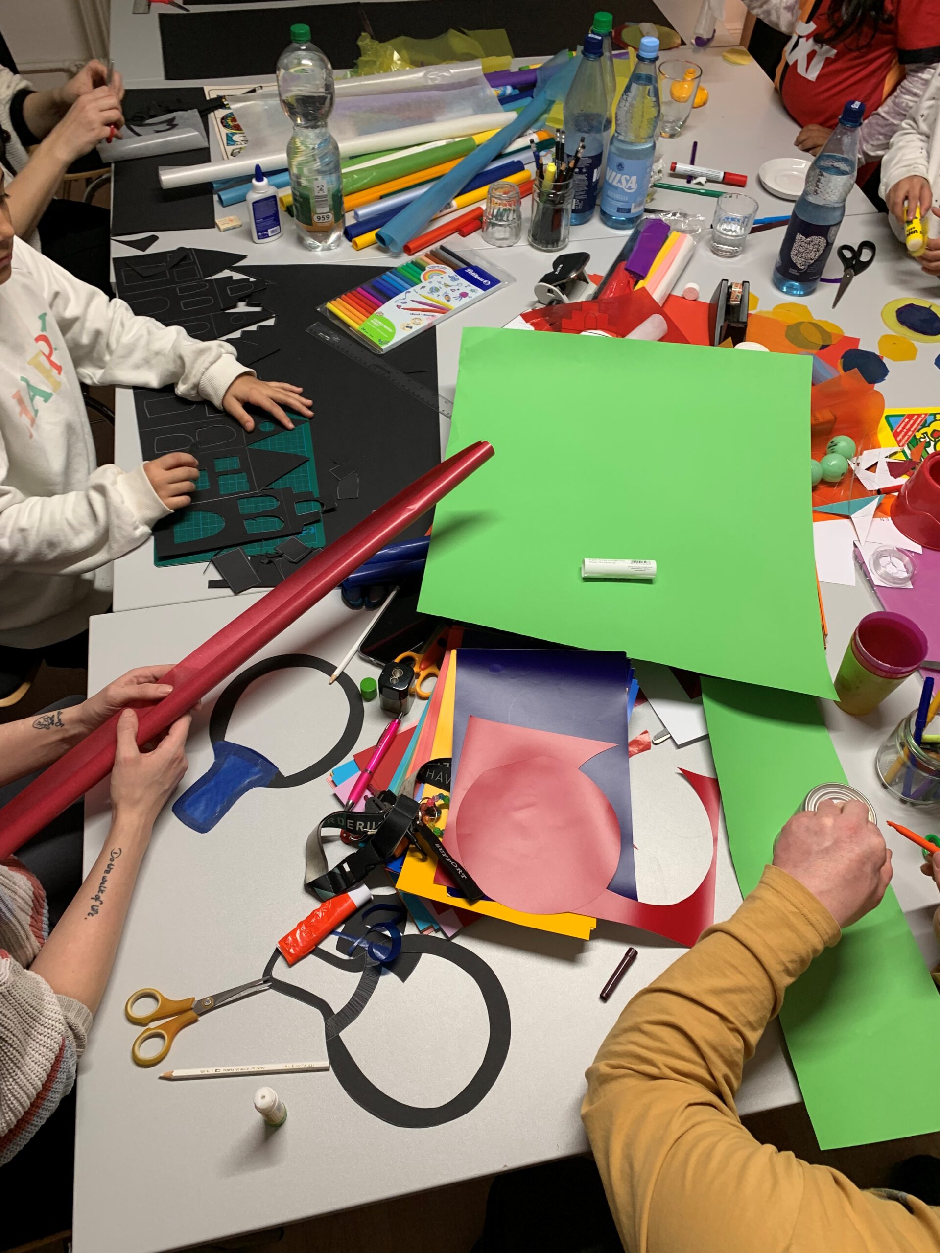 Viele verschiedene Bastelmaterialien liegen auf einem großen Tisch verstreut. Drumherum sitzen Kinder und Erwachsene, von denen man nur die Arme sieht. Alle sind am Basteln.