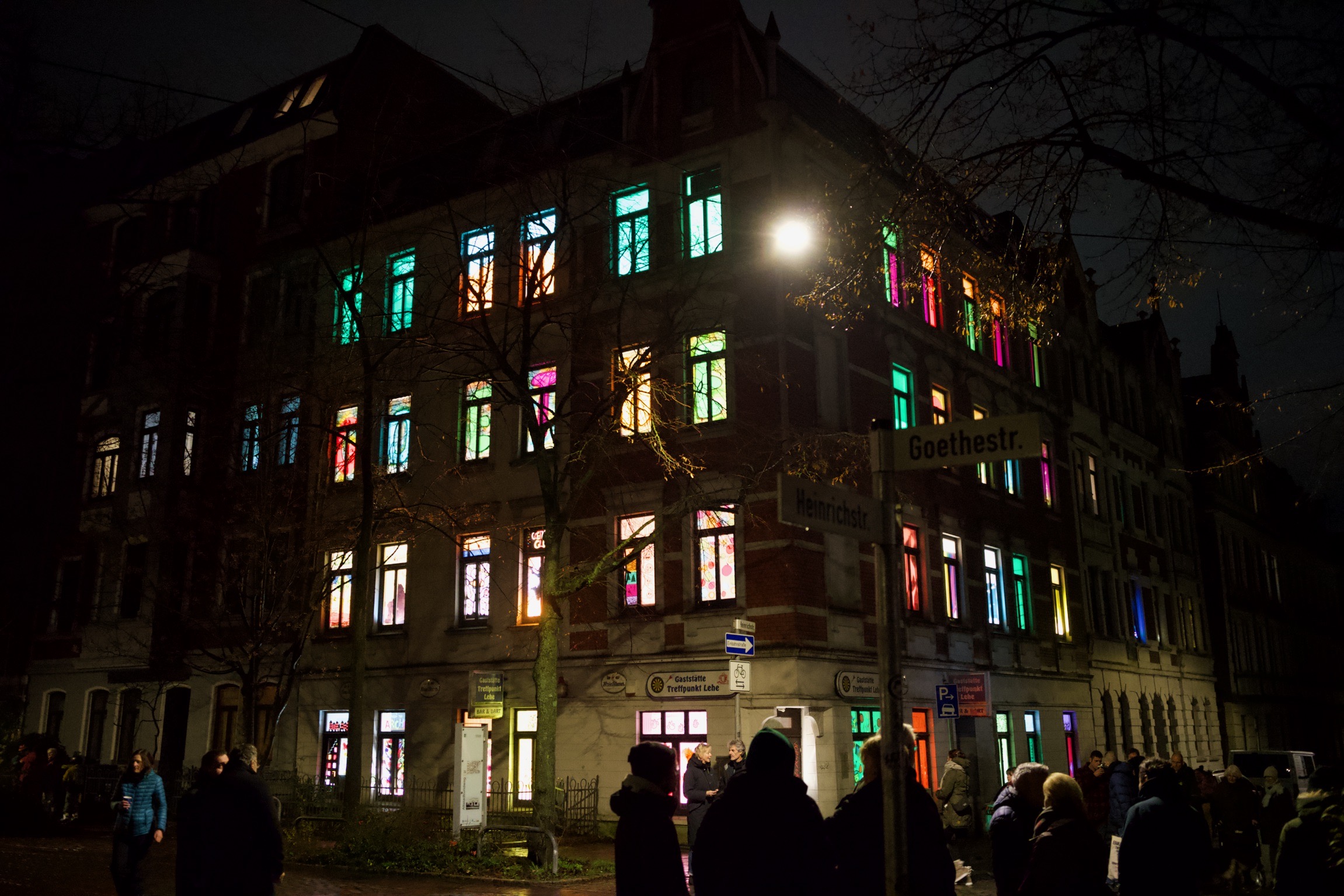 Ein vierstöckiges Altbauhaus in der Dunkelheit. Jedes der Fenster ist in einer anderen Farbe beleuchtet. Davor sind ein paar Menschen versammelt und schauen sich das Haus an.