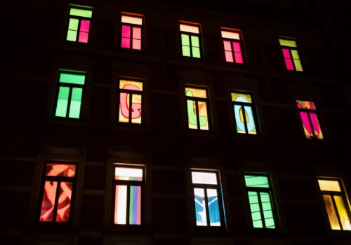 Fensterfront eines mehrstöckigen Altbauhauses in der Dunkelheit. Nur die beleuchteten Fenster sind zu erkennen. In jedem Fenster befindet sich eine andere bunte Gestaltung. In einer Reihe ergeben einzelne Buchstaben je Fenster, das Wort GLOW.