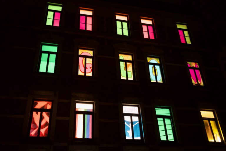 Fensterfront eines mehrstöckigen Altbauhauses in der Dunkelheit. Nur die beleuchteten Fenster sind zu erkennen. In jedem Fenster befindet sich eine andere bunte Gestaltung. In einer Reihe ergeben einzelne Buchstaben je Fenster, das Wort GLOW.