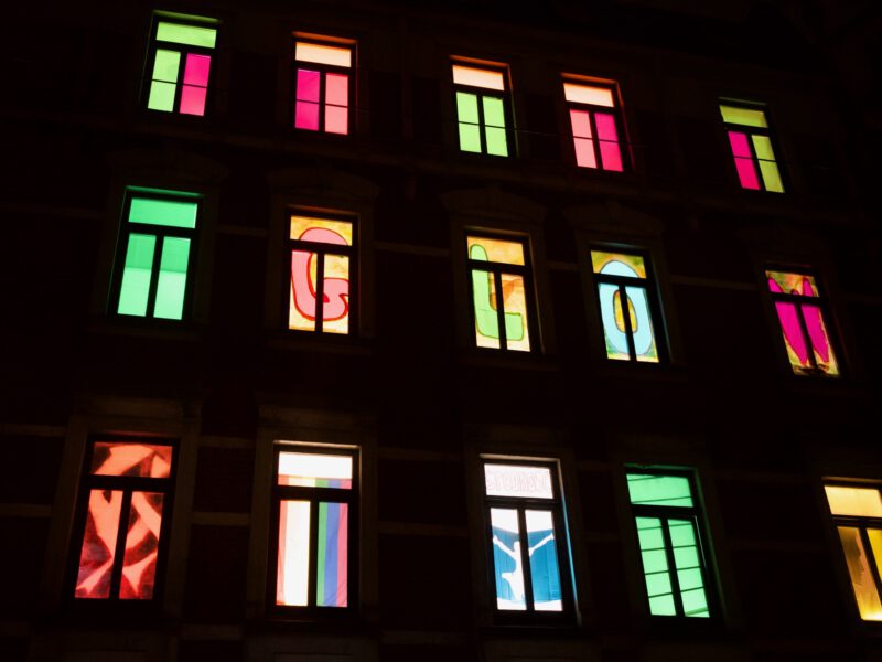 Fensterfront eines mehrstöckigen Altbauhauses in der Dunkelheit. Nur die beleuchteten Fenster sind zu erkennen. In jedem Fenster befindet sich eine andere bunte Gestaltung. In einer Reihe ergeben einzelne Buchstaben je Fenster, das Wort GLOW.